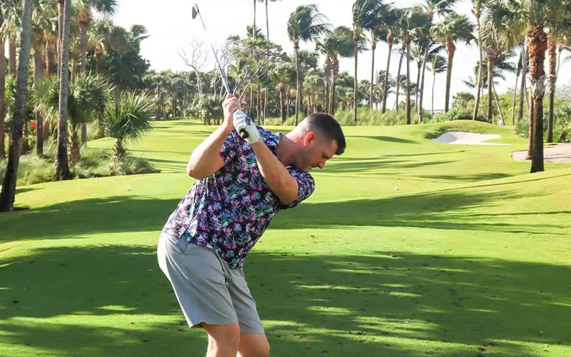 Man playing golf on lush green course with palm trees in the background.